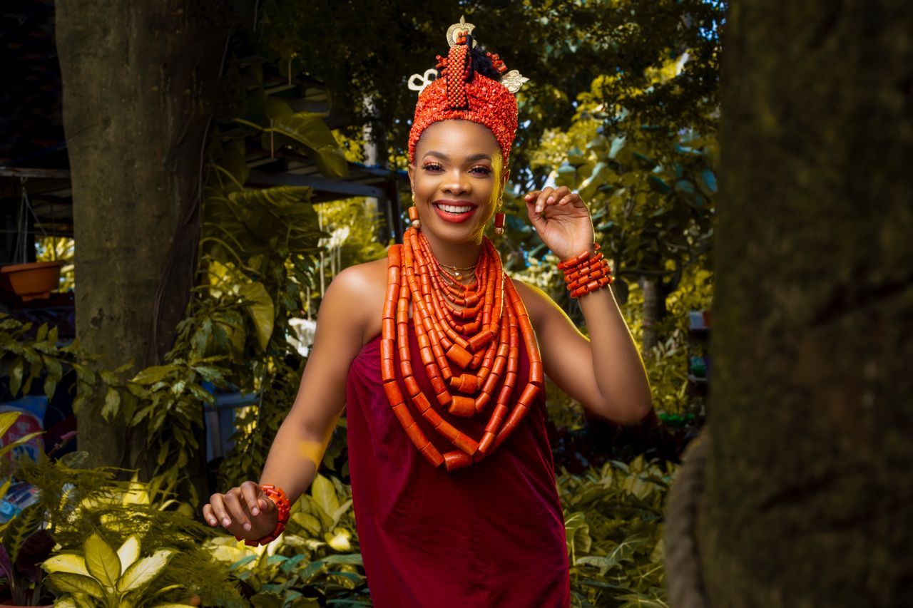 About Smiling woman in traditional attire with colorful beads in lush outdoor setting.
